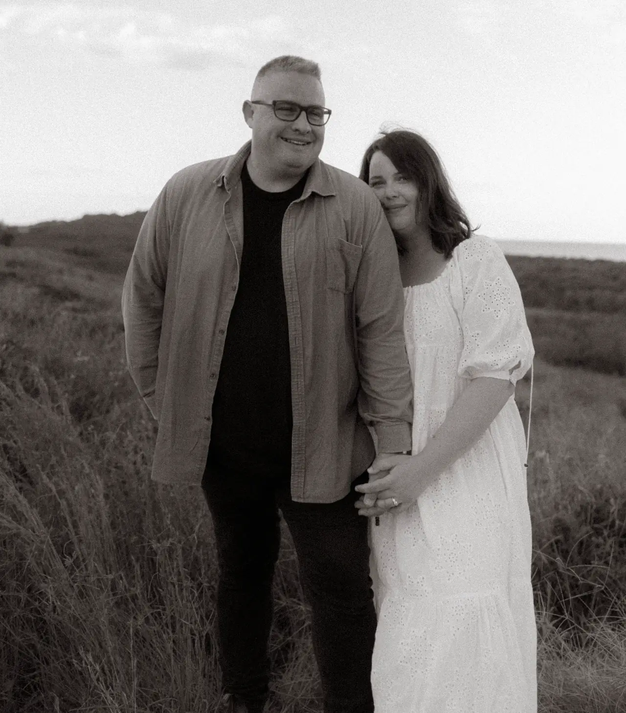 Black-and-white portrait of a couple standing together in coastal grass.